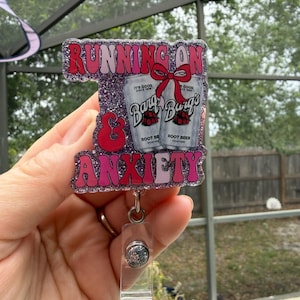 May include: A badge reel with the text "RUNNING ON & ANXIETY" in pink and red glitter. The badge features two root beer cans with a red bow. The reel is attached to a clear plastic holder.