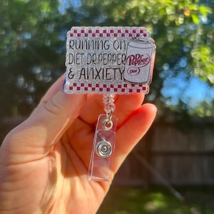 Può includere: Un portabadge rettangolare con la scritta "RUNNING ON DIET DR. PEPPER & ANXIETY" e una grafica di una lattina di Diet Dr. Pepper. Il badge ha un bordo glitterato e una clip a bobina retrattile in plastica trasparente.