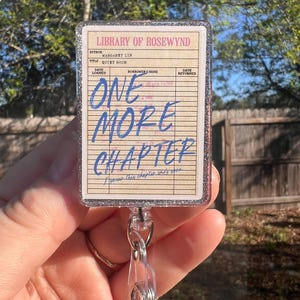 Op de afbeelding: Een badge reel met een bibliotheekkaartontwerp. De kaart leest "Library of Rosewynd" en "One More Chapter" in blauw schrift. De reel heeft een doorzichtig plastic intrekbaar koord en een zilveren glitterrand.