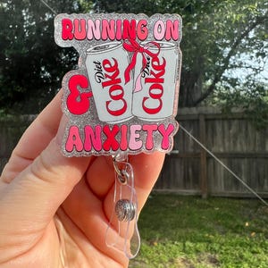 Puede incluir: Un portatarjetas rosa y plateado con el texto "RUNNING ON & ANXIETY". La insignia presenta dos latas de Coca-Cola Light atadas con un lazo rojo. El portatarjetas tiene un clip de plástico transparente y un cordón retráctil.