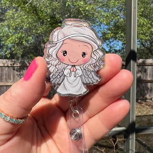 May include: A clear plastic badge holder with a glitter-covered image of a cartoon angel with a halo. The angel is wearing a white dress and has blonde hair. The badge holder has a retractable clip.