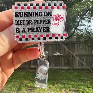 May include: A badge reel with the text "RUNNING ON DIET DR. PEPPER & A PRAYER". The badge has a glittery silver background with a red, black, and white checkered border. A Diet Dr. Pepper can graphic is on the right side.