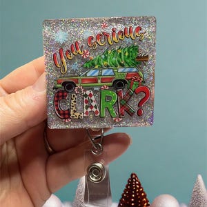 May include: A sparkly silver square badge holder with a Christmas-themed image of a car with a Christmas tree on top. The image says "You serious Clark?" in colorful letters.