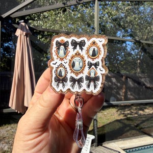 Può includere: Un portabadge quadrato con bordo glitterato dorato che presenta un motivo di fantasmi all'interno di cornici dorate ornate e fiocchi neri. Il design include piccoli pipistrelli neri sparsi tra le cornici. È fissato un cinturino in plastica trasparente.
