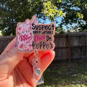 May include: A rose gold glitter badge reel with a Diet Dr. Pepper can design. The can is decorated with pink roses and the text "Suspect only drinks Diet Dr. Pepper". The reel has a clear plastic strap and a metal clip.