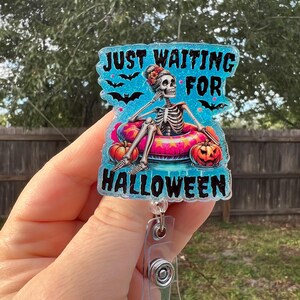 May include: A Halloween-themed badge reel with the text "JUST WAITING FOR HALLOWEEN." The badge features a skeleton relaxing in a pool float with pumpkins. The background is a light blue with black bats.