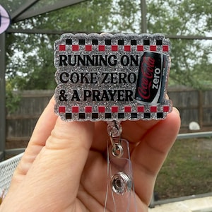 May include: A rectangular badge reel with the text "RUNNING ON COKE ZERO & A PRAYER". The badge reel has a black, red, and silver glitter design and features a Coca-Cola Zero can graphic.