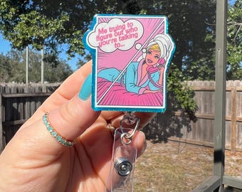 May include: A rectangular badge reel featuring a glittery blue border and a comic-style illustration of a blonde character in a blue robe and hair towel, holding a pink telephone. A speech bubble above reads, Me trying to figure out who you're talking to...