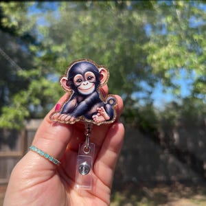 May include: A badge reel featuring a smiling cartoon chimpanzee with black fur and a gold glitter border. The badge reel has a clear plastic retractable cord and a silver-colored clip.