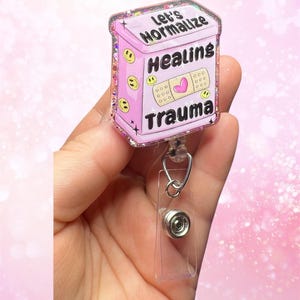 Peut inclure: Porte-badge en paillettes roses avec une image de dessin animé rose et blanche d'un flacon de pilules avec le texte "Let's Normalize Healing Trauma".