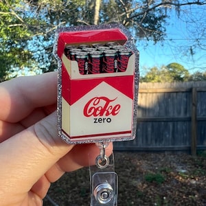 Op de afbeelding: Een novelty badge reel met een rood en wit ontwerp van een Coke Zero-blikjesdoos. De doos heeft het Coke Zero-logo in rode letters. De badge reel heeft een doorzichtige plastic clip en een zilveren glitterrand.