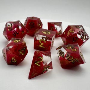 May include: Set of eight polyhedral dice in various shapes. The dice are transparent red with gold-colored numbers and internal red and gold marbling. Ideal for tabletop games like Dungeons and Dragons.