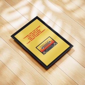 Peut inclure: Une affiche encadrée avec un fond jaune et un motif de cassette rétro. L'affiche présente le texte "THERE ARE THE HANDS THAT MADE US, AND THEN THERE ARE THE HANDS THAT GUIDE THEIR HANDS." Le cadre est noir.
