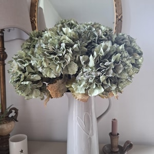 May include: A display of dried green hydrangeas in a white ceramic jug with a heart motif. A gold-framed mirror and a candle holder with a lit candle are in the background. A small white cup with a star is also visible.