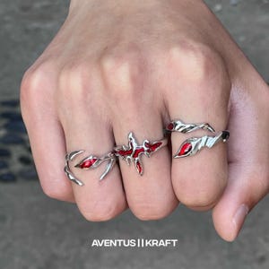 May include: Three silver rings with red enamel detailing are shown on a hand. The rings feature distinctive designs, including a flame-like shape, a spiky design, and a curved shape with a red stone. The text "AVENTUS || KRAFT" is at the bottom.