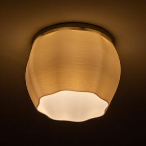 Può includere: Una plafoniera con un paralume strutturato bianco sporco. Il paralume ha una forma ondulata e organica e un interno bianco brillante. La lampada è posta su uno sfondo scuro, evidenziando il suo design.