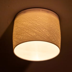 May include: A cylindrical ceiling light fitting with a woven, cream-coloured shade. The shade has a textured appearance, and the light emits a warm glow. The fitting is mounted on a white base.