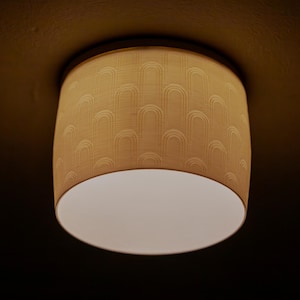以下が含まれることがあります： ベージュ色のドラム型シーリングライト。シェードにはアーチ型の形状が繰り返されるパターンがあります。ライトの下部は白く、点灯しています。ライトは濃い茶色の天井に取り付けられています。リビングルームに適しています。