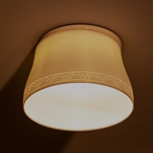 May include: A cream-colored, flush-mount ceiling light fixture. The light has a cylindrical shape with a decorative band of repeating semi-circles near the bottom. The light is illuminated, casting a warm glow.
