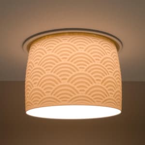 May include: A flush-mount ceiling light fixture with a cream-colored, cylindrical shade. The shade features a repeating, embossed wave pattern. The light is illuminated, casting a warm glow.