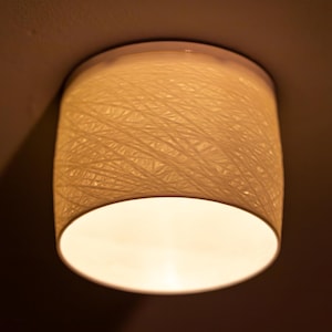 May include: A cylindrical ceiling light fixture with a woven, textured shade. The shade is a warm, off-white color, and the light emits a soft, inviting glow. The fixture is mounted on a white ceiling.