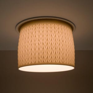 May include: A beige, cylindrical ceiling light fixture with a textured, woven pattern. The light has a circular base and a warm, soft glow emanating from the bottom. The fixture is mounted flush against the ceiling.