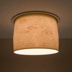 May include: A cylindrical ceiling light fixture with a textured, beige lampshade. The lampshade has a pattern of raised, organic shapes. The light fixture is mounted on a white ceiling and emits a soft, warm glow.
