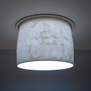May include: A white cylindrical ceiling light fixture with a floral pattern. The light is on, emitting a bright glow from the bottom. The fixture is mounted on a white ceiling.