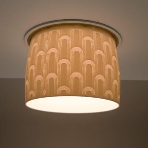 Könnte beinhalten: Eine beige Deckenleuchte mit einem sich wiederholenden Bogenmuster. Die Leuchte hat einen zylindrischen Schirm mit weißem Innenraum und eine kreisförmige Metallverkleidung. Das Licht ist eingeschaltet und beleuchtet den Raum.
