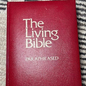 May include: A red hardcover book titled "The Living Bible" with the subtitle "Paraphrased" in white lettering. The book's cover is a solid, deep red color.