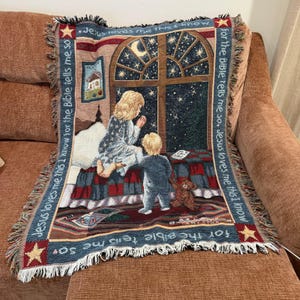 May include: A woven throw blanket featuring a scene of two children near a window at night. The border text reads, Jesus loves me this I know for the Bible tells me so. The image includes a crescent moon, stars, a teddy bear, and a framed picture.
