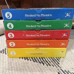 Puede incluir: Una pila de cinco cajas educativas rectangulares etiquetadas como Hooked On Phonics Learn to Read, dispuestas desde el nivel 5 en la parte superior hasta el nivel 1 en la inferior. Las cajas son azules, verdes, rojas, naranjas y amarillas.