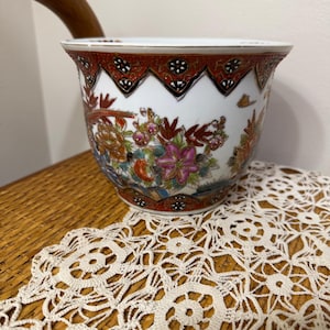 May include: A white porcelain bowl featuring a wide, flared rim decorated with a red, black, and gold geometric pattern. The exterior body displays a colourful floral motif with pink, orange, and green blossoms, accented by gold leaf detailing.