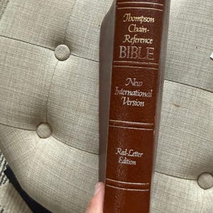 May include: A brown leather-bound Bible with gold lettering. The text on the spine reads "Thompson Chain-Reference BIBLE New International Version Red-Letter Edition". The book is held by a person.