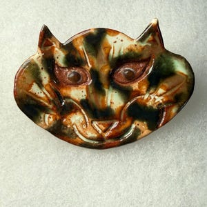 Pode incluir: Um prato de cerâmica em forma de gato com um design caprichoso. O prato apresenta um rosto com olhos castanhos, um nariz triangular e uma boca sorridente. A cobertura tem um padrão manchado de castanho, preto e verde.