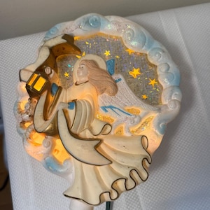 May include: A Christmas tree topper featuring an angel holding a lantern. The angel is dressed in a cream-colored gown with gold accents, surrounded by a blue and white cloud design. The background has a starry night scene with yellow stars.