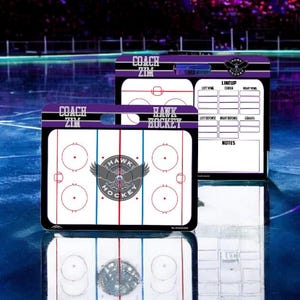 May include: Two dry-erase hockey coaching boards with a purple and black border. The top board has a lineup section and a notes section. The bottom board has a hockey rink diagram with a Hawk logo in the center. The text "COACH ZIM" is on both boards.