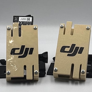 Pode incluir: Dois suportes de bateria DJI cor bege, com logótipo DJI preto e suportes de montagem pretos. Os suportes retangulares têm uma ranhura na parte superior e são fixados com quatro parafusos prateados. Uma bateria é visível num suporte.