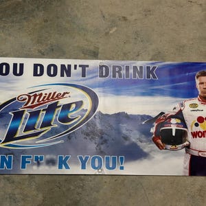 May include: Banner featuring the Miller Lite logo and the text "IF YOU DON'T DRINK THEN F**K YOU!". A man in a racing suit holds a helmet. The background shows a mountain scene.