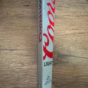 May include: A silver Coors Light beer tap handle featuring a prominent red Coors logo in raised lettering. The side displays the text Coors Light vertically. Near the base, a small triangle icon appears above the text Made with recycled cans.
