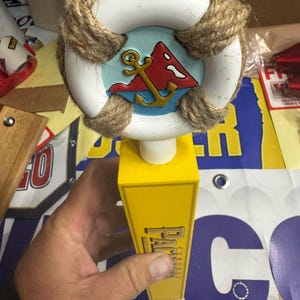 Puede incluir: Un tirador de grifo de temática náutica con un diseño de salvavidas blanco, un emblema de ancla roja y dorada sobre un fondo azul y detalles de cuerda. El tirador amarillo tiene el texto "PAL CERVEZA".