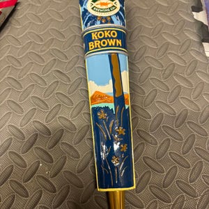May include: A Kona Brewing Co. Koko Brown beer tap handle featuring a blue, yellow, and white design. The top displays the brand logo with a lizard graphic, while the body depicts a landscape with a tree, flowers, and a mountain range.