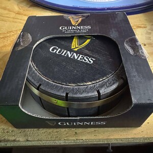 May include: A black and gold Guinness beer barrel shaped coaster in its original packaging. The packaging is black with the Guinness logo and text that reads "St. James's Gate Dublin, Ireland".