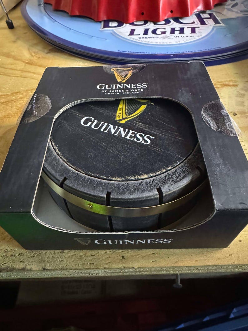 May include: A black and gold Guinness beer barrel shaped coaster in its original packaging. The packaging is black with the Guinness logo and text that reads "St. James's Gate Dublin, Ireland".