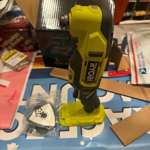May include: A lime green and dark grey Ryobi cordless oscillating multi-tool with a battery base. The tool features a vertical handle, a top-mounted metal attachment head, and a separate triangular sanding pad accessory enclosed in a clear plastic bag.