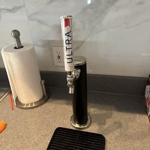 May include: A black beer tap with a silver spout and base, featuring the word "ULTRA" in white on the tap handle. A black drip tray sits below the tap. A roll of paper towels is to the left.