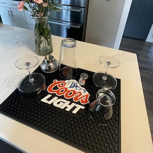 Może przedstawiać: Czarna mata barowa z logo Coors Light w kolorze czerwono-białym, otoczona kieliszkami do koktajli, shakerem i małym słoiczkiem. W tle wazon z różowymi kwiatami.