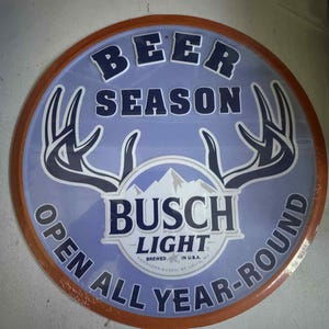 以下が含まれることがあります： 薄い青色の背景と茶色の木製フレームの丸い看板。「BEER SEASON」と「OPEN ALL YEAR-ROUND」の文字と中央に「BUSCH LIGHT」のロゴが入っています。鹿の角のデザインも特徴です。