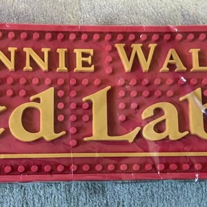 May include: A red bar mat with the words "JOHNNIE WALKER Red Label" in gold lettering. The mat has a textured surface with small, raised dots and is rectangular in shape.