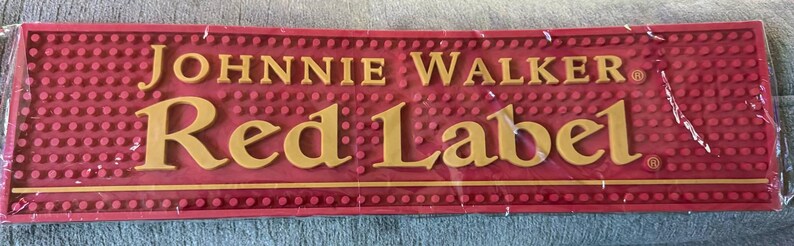 May include: A red bar mat with the words "JOHNNIE WALKER Red Label" in gold lettering. The mat has a textured surface with small, raised dots and is rectangular in shape.